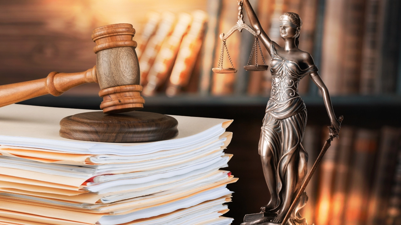 Ein Richterhammer liegt auf einem Aktenstapel, daneben steht eine Statue von Justizia, im Hintergrund sieht man unscharfe Bücher