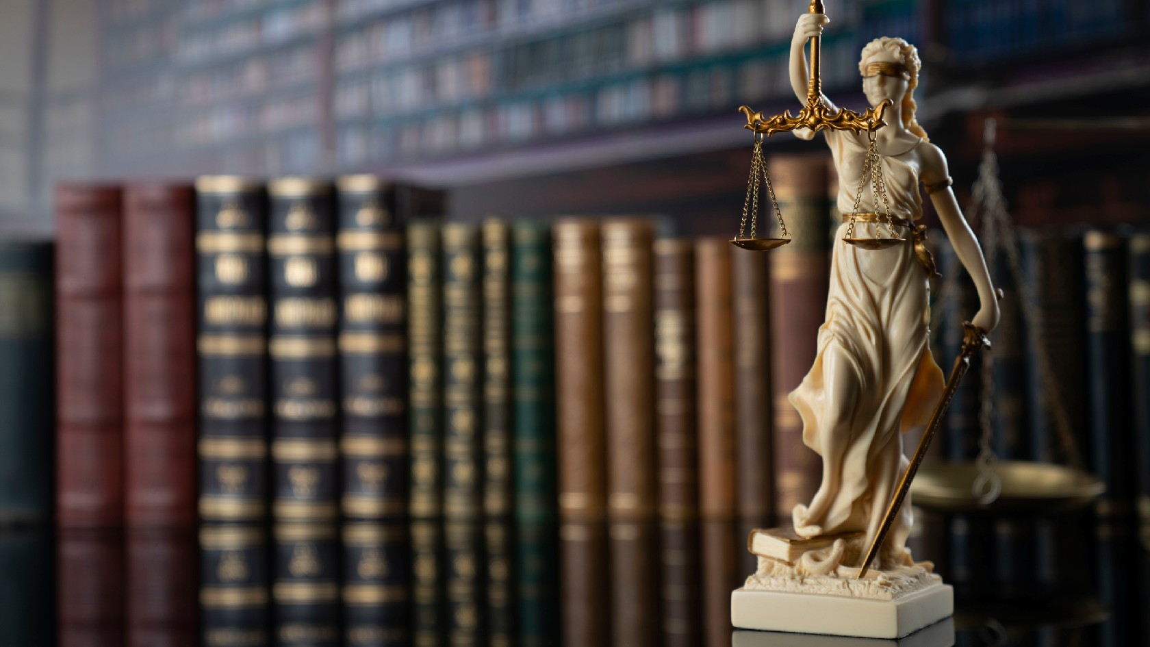 Eine Miniaturstatue von Justizia, welche eine Waage hält steht vor einer Reihe von Rechtsbüchern im Hintergrund
